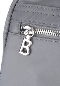 Borsa in tessuto grigia con zip argentata e un ciondolo a forma di lettera "B" attaccato, che mostra un motivo testurizzato e cuciture finemente realizzate.