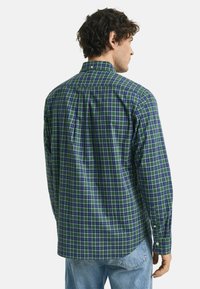 Mann, der von hinten zu sehen ist, trägt ein grün-navy kariertes Hemd mit Knöpfen und hellblaue Jeans vor einem schlichten Hintergrund.