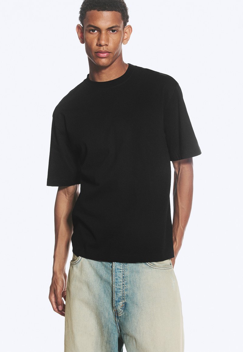 Man draagt een eenvoudige zwarte korte mouwen T-shirt en lichtblauwe spijkerbroek, staand tegen een effen witte achtergrond.