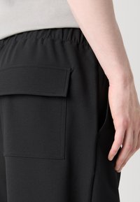Svart joggebukse med elastisk midje, glatt stofftekstur, sidelommer og en rektangulær lappelomme bak.