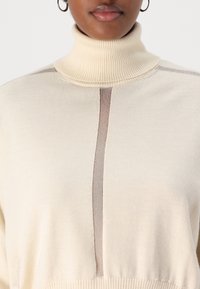 Krēmkrāsas augstais apkakles džemperis ar teksturētu virsmu, kam centrā ir smilšu krāsas svītra un ribota apdare apkakles un apakšmalas daļā.