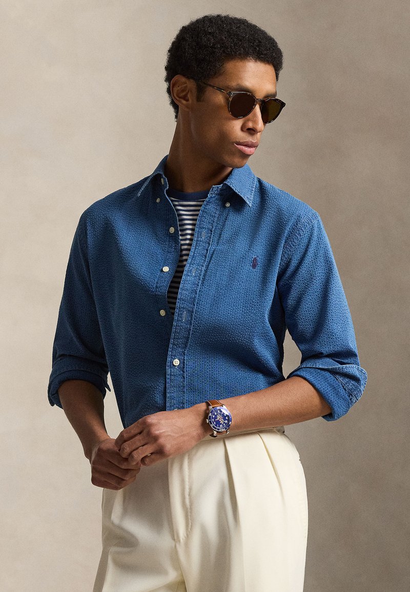 Mees kannab tumedaid päikeseprille, sinist tekstuuriga särki triibulise T-särgi peal, valgeid volditud pükse ja sinise kellakettaga pruuni nahkrihmaga kella.