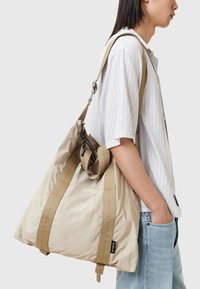 Bolso tote de nailon beige con correas anchas de lona y costuras rojas en contraste, que presenta un clip desmontable y una forma triangular aerodinámica.