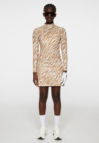 Långärmad, figurnära klänning i tan med ett vitt vågymönster; tillverkad av stretchmaterial. Matchas med solglasögon samt vita sportstrumpor och skor.