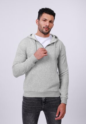 Hombre con cabello rizado corto vestido con una sudadera gris con una camiseta blanca debajo, sosteniendo la cremallera de la sudadera con una mano, de pie.