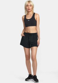 Sort sportssnor med hvidt logo, parret med sorte shorts med elastisk talje og lommer samt sorte træningssko. Neutral baggrund.
