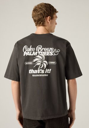 Jeune homme vu de dos portant un T-shirt sombre avec un texte blanc et un graphisme de palmier indiquant "Calm Breeze Palm Trees, that's it!"