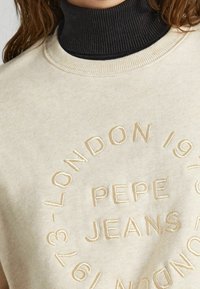 Crème sweatshirt met een zwart geribd coltrui, voorzien van verhoogde beige borduring van "PEPE JEANS LONDON 1973" in een cirkelontwerp.