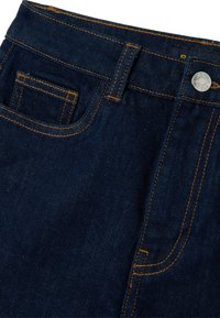Jeans de mezclilla azul oscuro con costuras naranjas, un bolsillo frontal para monedas, passadores para cinturón y cierre de botón plateado.