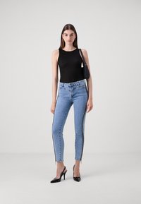 Fekete bordázott ujjatlan top, világoskék skinny farmernadrággal, oldalsó cipzárakkal és rojtos szegélyekkel. A modellen fekete pántos magassarkú cipő van.