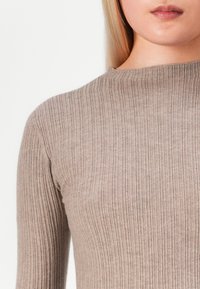 Kobieta nosząca beżowy sweter o długim rękawie z prążkowanej dzianiny i wysokim okrągłym dekoltem, ukazująca górną część tułowia i częściowo twarz.