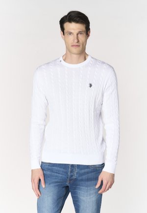 Uomo che indossa un maglione bianco a trecce e jeans blu, in piedi davanti a uno sfondo chiaro e uniforme, rivolto in avanti.