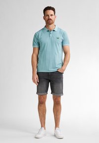 Světle modré polo tričko s límcem a malým logem na prsou, v kombinaci s šedými denimovými šortkami. Model má na sobě bílé tenisky. Jednoduché pozadí.