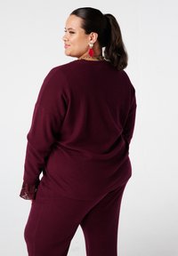 Pull en maille côtelée bordeaux à coupe décontractée, avec des accents en dentelle sur les manches. Assorti à un pantalon pour un look coordonné.