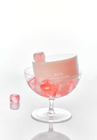 Récipient circulaire rose contenant des patches pour les yeux dans un bol en verre transparent rempli de cubes de gel rose translucides, reflétant la lumière sur un fond blanc.