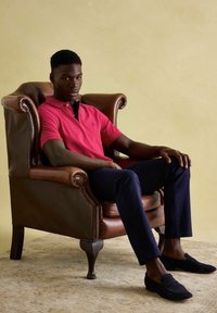 Mladý muž v červeném polu košili a námořnických kalhotách sedí uvolněně v hnědé kožené křesle na béžovém pozadí.