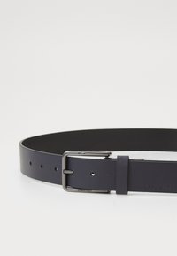 Mørkeblå læderbælte med en glat overflade, udstyret med en rektangulær sort metalspænde og flere justeringshuller til tilpasning.