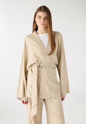 Giovane donna con capelli lunghi indossa una giacca beige con cintura, rifiniture in pizzo e pantaloni coordinati, in piedi su sfondo bianco.