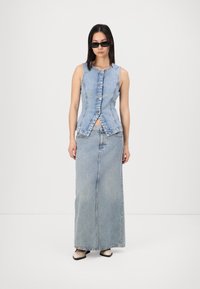 Lichtblauw denimvest met een knoopsluiting aan de voorkant, gecombineerd met een lange, bijpassende denim rok. Gelaagd met zwarte rechthoekige zonnebril en beige schoenen.