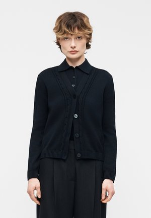 Young person with short wavy hair wearing a black buttoned cardigan over a black collared shirt and black pants against a plain background.