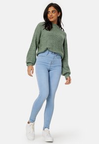 Zielony sweter z dzianiny z długimi rękawami, w połączeniu z jasnoniebieskimi, obcisłymi dżinsami z wysokim stanem i białymi trampkami, na jednolitym tle.