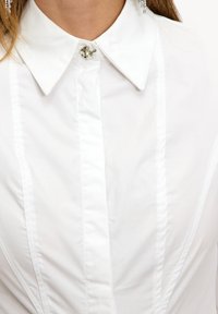 Blusa blanca con cuello puntiagudo, botón decorativo plateado, pliegues verticales y textura de tela suave.