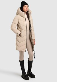 Beige pufferjack met een hoge kraag, een ruitvormig quiltontwerp en een ritssluiting. Draagt een aansluitende beige legging en zwarte kniehoge laarzen.