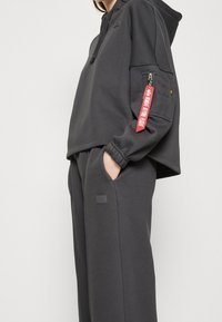 Ciemnoszara bluza z kapturem z zamykaną na zamek kieszenią na rękawie i czerwoną metką. Miękki materiał, luźny krój oraz dopasowane, obszerne spodnie dresowe z naszywką z logo.