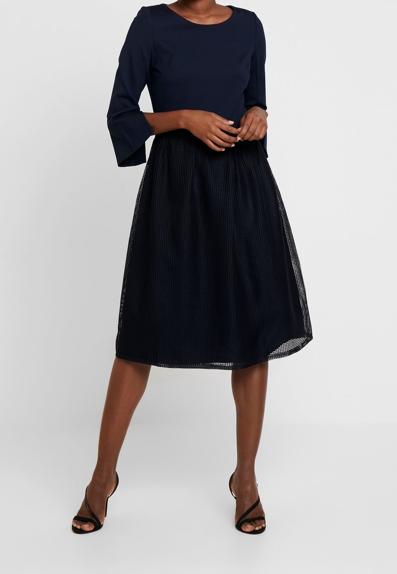 Navy blue dress with a fitted top and flared, knee-length skirt made of textured fabric. Features three-quarter sleeves and sheer overlay.