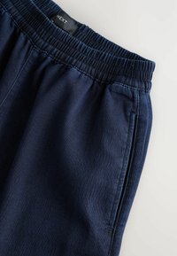 Pantaloni jogger cu talie elastică, de culoare albastru marin, fabricați dintr-un material texturat. Au buzunare laterale și cusături vizibile pe talie.