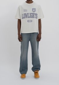 Bílý oversized bavlněný tričko s grafikou "LOWLIGHTS 2017" v šedé barvě. V kombinaci se světle modrými džíny a hnědými pracovními botami.