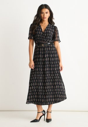 Woman wearing a black midi dress with small diamond patterns, short sheer sleeves, a black belt, and black high-heeled shoes, standing against white wall.