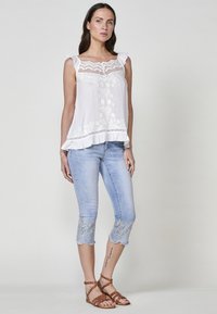 Haut sans manches blanc avec des détails en dentelle et des bords froncés, associé à un jean capri bleu clair avec des ourlets brodés et des sandales brunes à lanières.