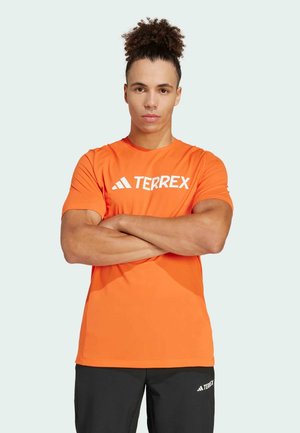 Narančasta majica s kratkim rukavima, sportski kroj, s TERREX logotipom u bijeloj boji. Glatka tekstura, dizajn s okruglim izrezom, uparen s crnim hlačama.