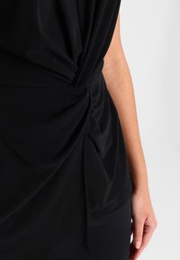 Top negro con detalle de nudo en el lateral, tela suave, diseño drapeado y textura lisa, combinado con pantalones negros ajustados.