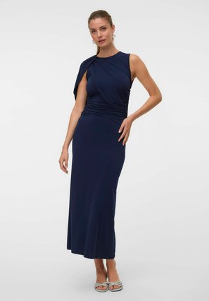 Femme en robe longue bleu marine sans manches avec des détails drapés, debout, une main sur la hanche et portant des talons ouverts clairs.