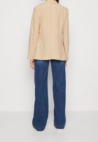 Person som bär beige kavaj med randig mönstring och vida blå jeans, står mot en enfärgad vit bakgrund, vänd bort.