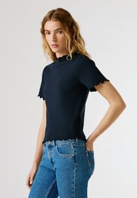 Μπλε σκούρο ριπ T-shirt με υψηλό λαιμόκοψη και κυματιστές άκρες· κοντά μανίκια· εφαρμοστή γραμμή· διακοσμημένο με μικρό λογότυπο στο στήθος.