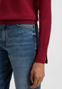Maglione a maglia color granata con bordi a coste, indossato sopra dei jeans di denim blu. Il maglione presenta delle fessure laterali all'orlo per un tocco di design in più.