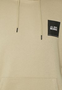 Sudadera beige con un bolsillo frontal, cordones y un parche rectangular negro con texto blanco. Textura suave y diseño casual.