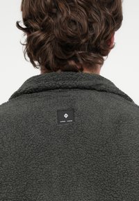 Antraciet fleecejack met een zachte structuur, voorzien van een sjaalkraag en een zwart label met witte tekst aan de achterkant.