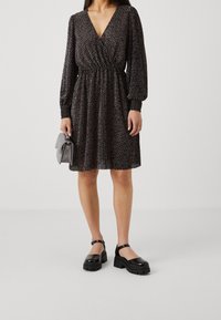Schwarzes Kleid mit V-Ausschnitt, langen Ärmeln und elastischem Taillenbund, mit einem weißen Pünktchenmuster; kombiniert mit schwarzen Plateauschuhen und einer grauen Handtasche.