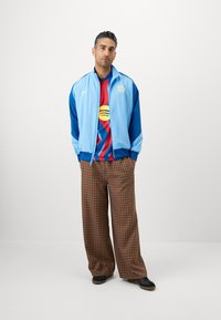 Casaco de zíper azul claro com detalhes azuis, combinado com uma t-shirt listrada em vermelho e azul com um logo circular. Calças largas de xadrez castanho.