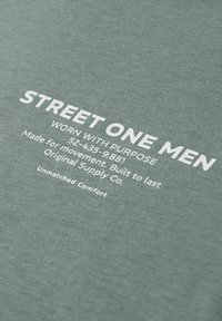 Zielona bawełniana koszulka z białym nadrukiem. Nadruk zawiera nazwę marki, cel oraz przekaz o komforcie, z gładkim, fakturowanym wykończeniem.