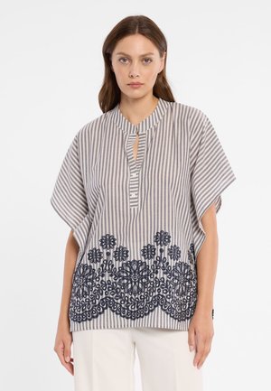 Vrouw draagt een losse blouse met korte mouwen, gestreept en met een mandarijnkraag, voorzien van marineblauwe bloemrijke borduursels aan de onderkant aan de voorkant.