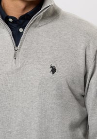 Man wearing light gray quarter-zip sweater over dark navy collared shirt, sweater featuring small black embroidered logo on chest.