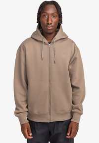 Brown zip-up hoodie with a drawstring hood, kangaroo pockets, and ribbed cuffs. Features subtle branding on the chest and a relaxed fit.