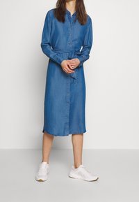 Abito camicia in denim di un blu medio, con chiusura a bottoni sul davanti, maniche lunghe, vita regolabile con cintura e orlo a lunghezza ginocchio.