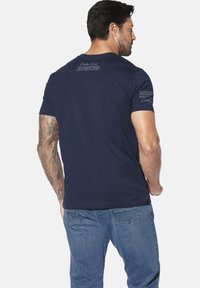 Navy blue cotton T-shirt with short sleeves, featuring logo text on back and sleeve. Simple design, relaxed fit, smooth texture.