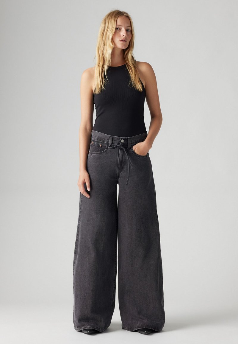 Schwarz denim jeans mit weitem Bein und hoher Taille, detaillierten Nähten und einem Zipper. Kombiniert mit einem enganliegenden schwarzen Tanktop.